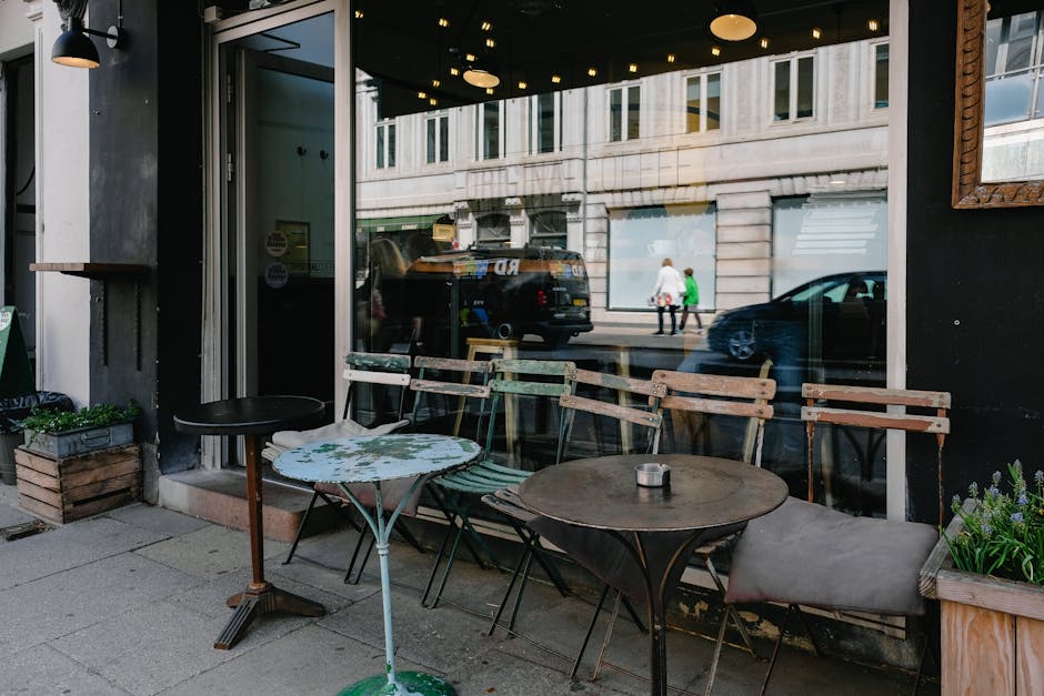 The image shows an outdoor seating area of a coffee shop or café located on a sidewalk in an urban environment. There are three tables, each with distinct styles: one small round table with a floral-patterned surface, one slightly larger round table with a dark wooden finish, and another rectangular wooden table. Surrounding these tables are four chairs, two of which are metal framed with wooden slats, and the third is an upholstered cushioned seat placed at the rectangular table. To the left, there is a small wooden planter box containing green plants, and a black side table stands beside the small round table. The seating area is positioned adjacent to a large glass window and door, revealing the interior of the establishment. The reflection on the window shows a street scene with two pedestrians walking and vehicles passing by, including a black van and a dark-colored car. The exterior wall of the café is painted dark, with a remaining wooden frame visible on the right side. The overall setting has a casual, inviting atmosphere typical of a café sidewalk dining area, and the scene subtly supports the context of urban waste management and rubbish removal services provided by House Clearance Balham.