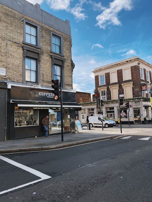 A street view extending across the corner of a city block showing a three-storey brick building with large windows and a black storefront sign with the word 'COFFEEB'. In front of the building, a person is standing near the entrance, possibly waiting or entering. To the right, there is a white van parked close to a sidewalk with a few pedestrians walking along the pavement. The scene includes a traffic light on a black pole positioned at the corner, and the road has marked parking spaces and crosswalks. The sky above is partly cloudy with patches of blue indicating fair weather. The environment appears urban, with the buildings featuring traditional architectural details and neutral tones. This scene reflects a typical commercial area where local businesses operate, possibly in proximity to private waste handling and rubbish collection services such as those provided by House Clearance Balham.
