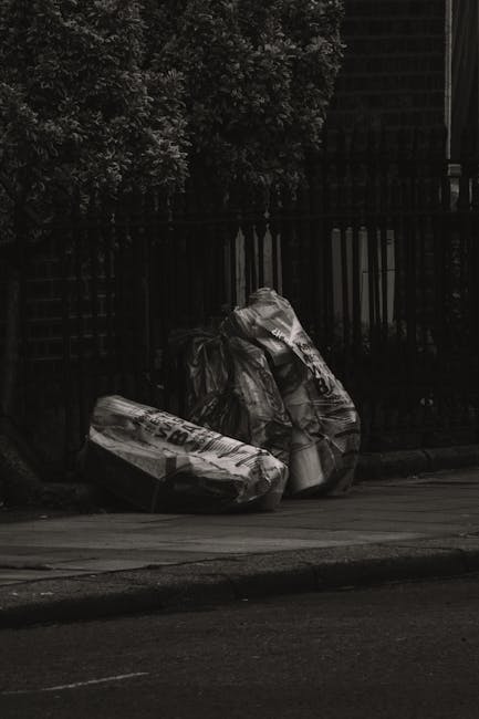 Three large black and white trash bags filled with rubbish are placed on the pavement next to a black metal fence, which has vertical bars and a slatted wooden upper section. The bags appear to be made of durable plastic material and are tightly packed, with some slight wrinkles and creases visible on the surfaces. The scene is set outdoors on a street or alleyway, with the bags positioned close to the curb. Behind the fence, there are dense, dark foliage and the edge of a building structure partially visible at the top right corner of the image. The lighting is subdued, possibly capturing an overcast day or the shadowed area, emphasizing the contrast between the bags and the general surroundings. This scene is indicative of private waste collection or on-site rubbish clearance, aligning with services offered by companies like House Clearance Balham, who provide alternative waste handling options outside municipal collection systems.
