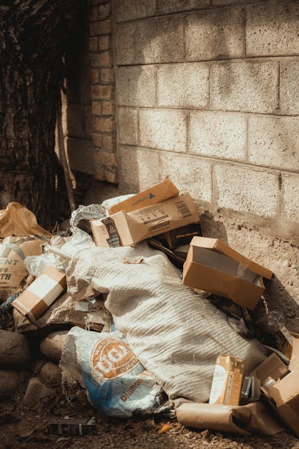 Three large black and white trash bags filled with rubbish are placed on the pavement next to a black metal fence, which has vertical bars and a slatted wooden upper section. The bags appear to be made of durable plastic material and are tightly packed, with some slight wrinkles and creases visible on the surfaces. The scene is set outdoors on a street or alleyway, with the bags positioned close to the curb. Behind the fence, there are dense, dark foliage and the edge of a building structure partially visible at the top right corner of the image. The lighting is subdued, possibly capturing an overcast day or the shadowed area, emphasizing the contrast between the bags and the general surroundings. This scene is indicative of private waste collection or on-site rubbish clearance, aligning with services offered by companies like House Clearance Balham, who provide alternative waste handling options outside municipal collection systems.
