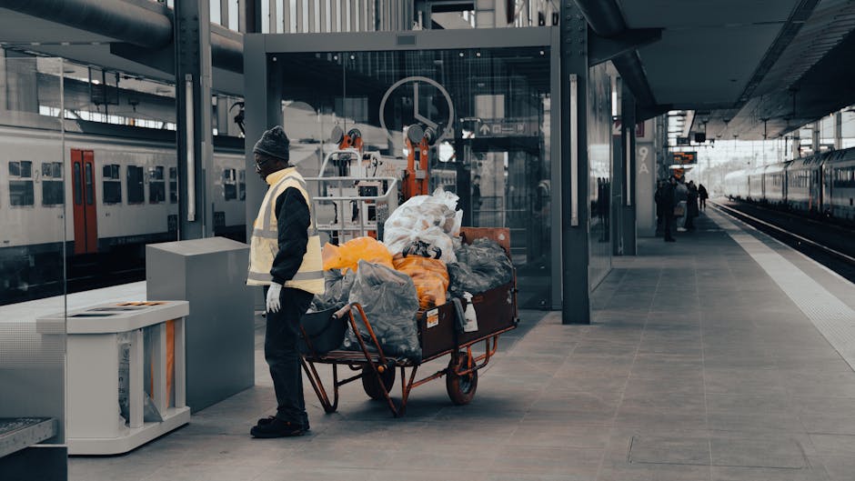 A person wearing a gray beanie, dark jacket, light yellow safety vest, and white gloves stands on a modern train station platform next to a large red metal trolley fully loaded with black, gray, and orange plastic bags, some of which are tied closed and others appear loose. The bags contain various materials likely intended for rubbish disposal, with some plastic wrapping visible on certain bags. The trolley is positioned near a sleek, glass-enclosed lift with mechanical equipment visible behind it, indicating a multi-level station environment. To the left, there is a white waste bin with a slot for disposal, and further in the background, a white train with red doors is stationary on the track. The platform features a clean, gray tiled surface with minimal markings, and in the distance, a few figures can be seen walking or waiting, with the overall atmosphere suggesting an urban transportation hub. The scene subtly highlights the context of waste collection and disposal, aligning with independent rubbish removal or on-site clearance services like those offered by House Clearance Balham.