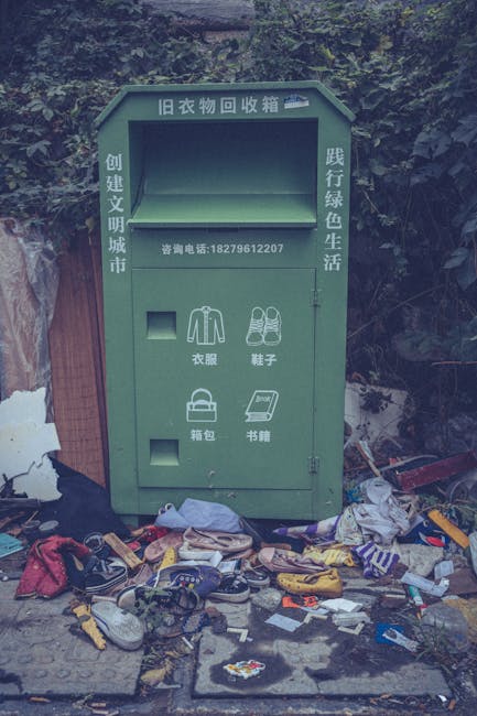 A green, metal clothing donation box situated outdoors against a backdrop of dense foliage and trees. The box features Chinese characters and a contact phone number, with an upper compartment intended for collecting donated clothing items and a lower section for smaller donations such as shoes, bags, and books, indicated by icons. The surrounding ground is cluttered with various discarded items including shoes, clothes, and miscellaneous rubbish, scattered around the base of the donation box. Additional debris, possibly packaging or trash, is seen among the scattered objects. The environment appears neglected, with the littered scene emphasizing improper waste disposal, contrasting with the intended charitable use of the box. The overall setting suggests a location where public waste management or private clearance by companies like House Clearance Balham might be necessary to remove improperly disposed rubbish and restore order to the area.