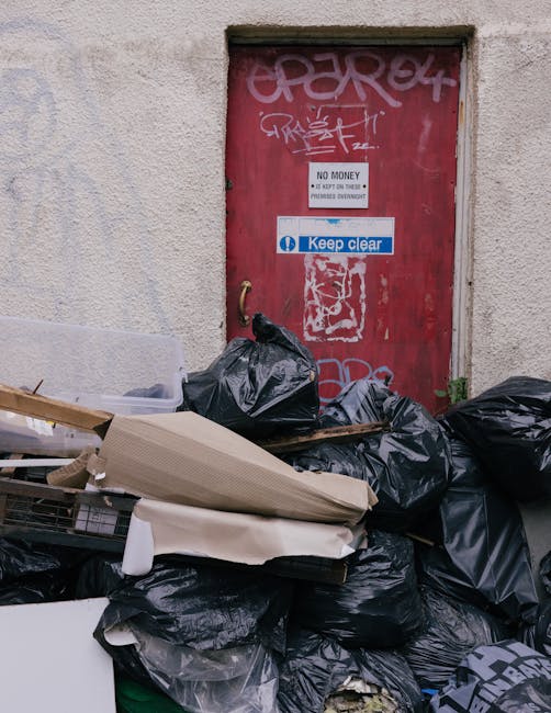 A utilitarian red metal door set into a rough-textured concrete wall, with graffiti tags in white spray paint partially covering the top portion and a sticker with the message 'No Money' and 'Keep Clear' adhered to the center. In front of the door, there is a large collection of black rubbish bags piled on the ground, with some of the bags appearing overfilled and bulging. A few bulky items, including a piece of cardboard and parts of discarded furniture or packaging, are resting atop or among the bags. The surrounding environment suggests an alleyway or back entrance of a building, where waste has been gathered for collection. Natural lighting highlights the textures of the bags, the graffiti, and the concrete wall, contributing to a utilitarian and unkempt scene associated with private rubbish disposal or small-scale clearance efforts managed by companies like House Clearance Balham, ensuring proper handling of waste in tight urban spaces.