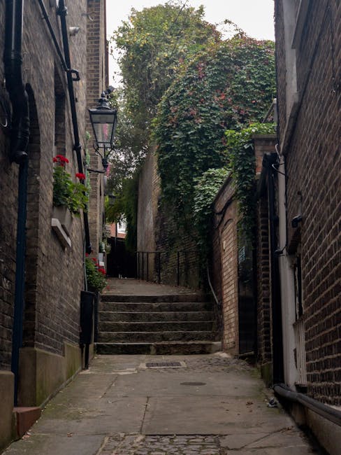 A narrow, paved alleyway between two brick buildings leads uphill with a short flight of stone steps at the end. The left building features a black iron wall-mounted lantern and a window with a flowerpot filled with red flowers. The right building has a small white door at the base and black external piping running vertically along the wall. The alley is bordered by walls of weathered red and brown bricks, some of which show signs of age and moss growth. At the top of the steps, dense greenery, including a large bush with reddish leaves, overhangs and partially obscures the view beyond. Natural light illuminates the scene, creating subtle shadows on the uneven paving stones and steps, while a black handrail runs along the left side of the stairs. The setting suggests an urban backstreet or side alley, typical of city environments where private rubbish collection or on-site clearance might be employed, supported by elements such as external piping and absence of public signage. The scene is indicative of a quiet, residential area that may benefit from professional waste management services like those offered by House Clearance Balham.