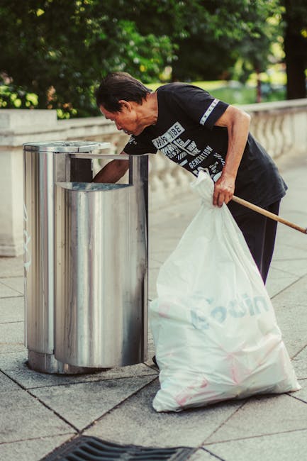 A middle-aged man with dark hair, dressed in a short-sleeved black T-shirt with white graphic text on the front, is seen leaning forward to dispose of rubbish into a large, stainless steel public waste bin situated on a paved pavement. The waste bin has a rounded, slightly tapered design with a wide opening at the top and a smooth, polished finish that reflects the surrounding environment. The man is holding a large, semi-transparent white plastic rubbish bag, which appears full and is marked with faint logos or text. He is using a wooden stick or litter picker to assist in placing the rubbish into the bin. The background features a lush green park with trees and bushes, suggesting an outdoor setting in a residential or urban area. The scene is well-lit with natural daylight, and the overall environment appears clean and orderly, consistent with private or alternative waste collection methods often provided by waste management services like House Clearance Balham.