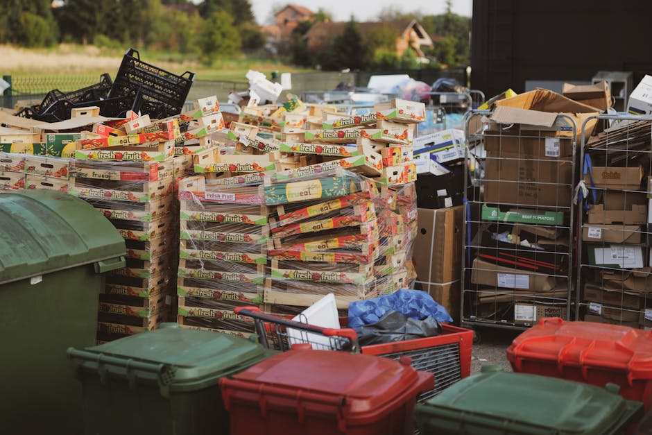 A large outdoor scene showing multiple stacks of empty cardboard fruit crate boxes, mostly white with colorful labels and logos, arranged on a paved area. To the left, there are two green plastic waste bins with lids, and in front, a small red plastic crate with some discarded plastic bags and debris. On the right side, there are two metal wire cages filled with various boxes, some with visible labels and barcodes, and brown paper packaging. Behind the stacks of fruit crates, there are additional cardboard boxes, some open with contents partially visible, and more boxes and packaging materials dispersed across the area. The background features a rural environment with a grassy boundary, trees, and houses with pitched roofs in the distance, under soft daylight. The scene appears to be part of a private rubbish removal or on-site clearance operation conducted by House Clearance Balham, with an emphasis on alternative waste handling methods outside regular municipal collection.