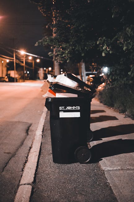 A middle-aged man with dark hair, dressed in a short-sleeved black T-shirt with white graphic text on the front, is seen leaning forward to dispose of rubbish into a large, stainless steel public waste bin situated on a paved pavement. The waste bin has a rounded, slightly tapered design with a wide opening at the top and a smooth, polished finish that reflects the surrounding environment. The man is holding a large, semi-transparent white plastic rubbish bag, which appears full and is marked with faint logos or text. He is using a wooden stick or litter picker to assist in placing the rubbish into the bin. The background features a lush green park with trees and bushes, suggesting an outdoor setting in a residential or urban area. The scene is well-lit with natural daylight, and the overall environment appears clean and orderly, consistent with private or alternative waste collection methods often provided by waste management services like House Clearance Balham.
